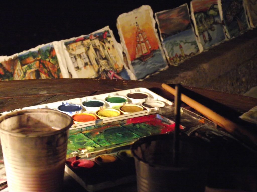 A palette of watercolors on the table and paintings drying in the background