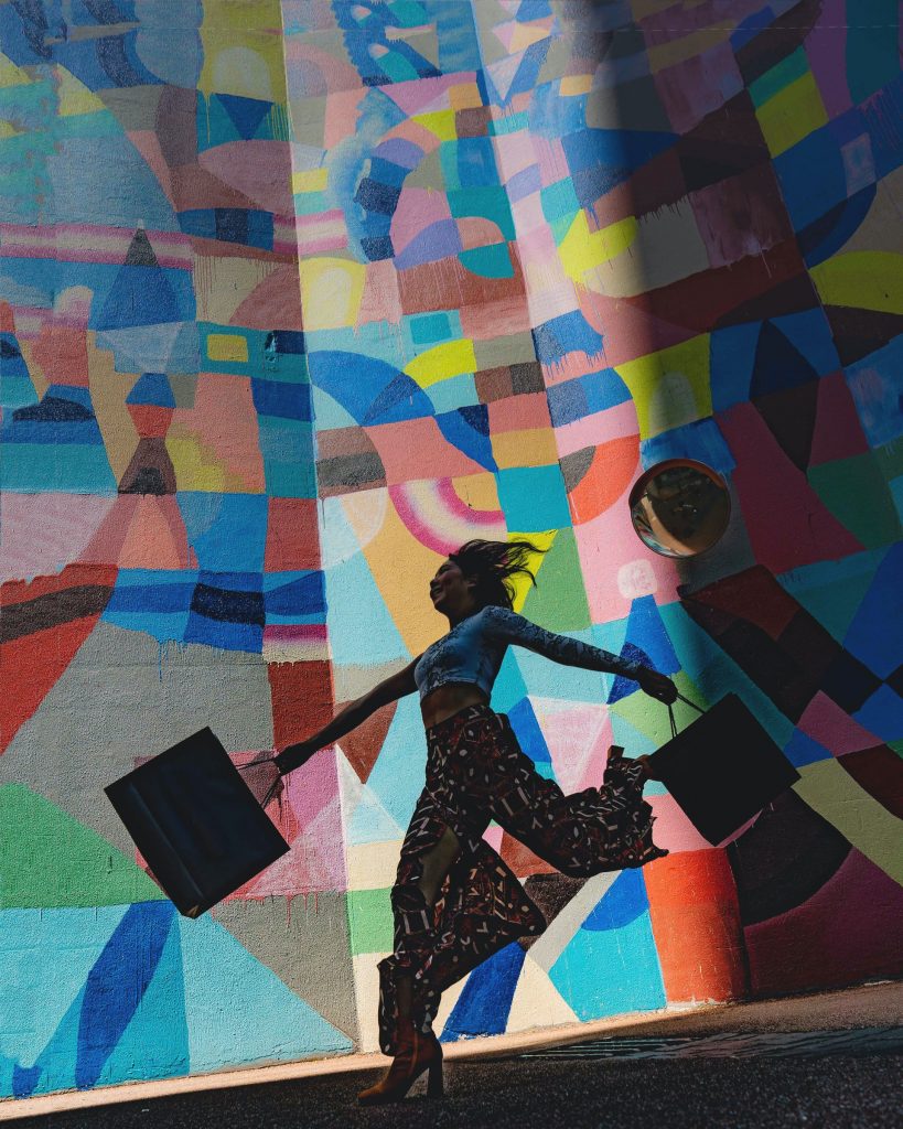Woman carrying bags jumping for joy in front of a colorful mural